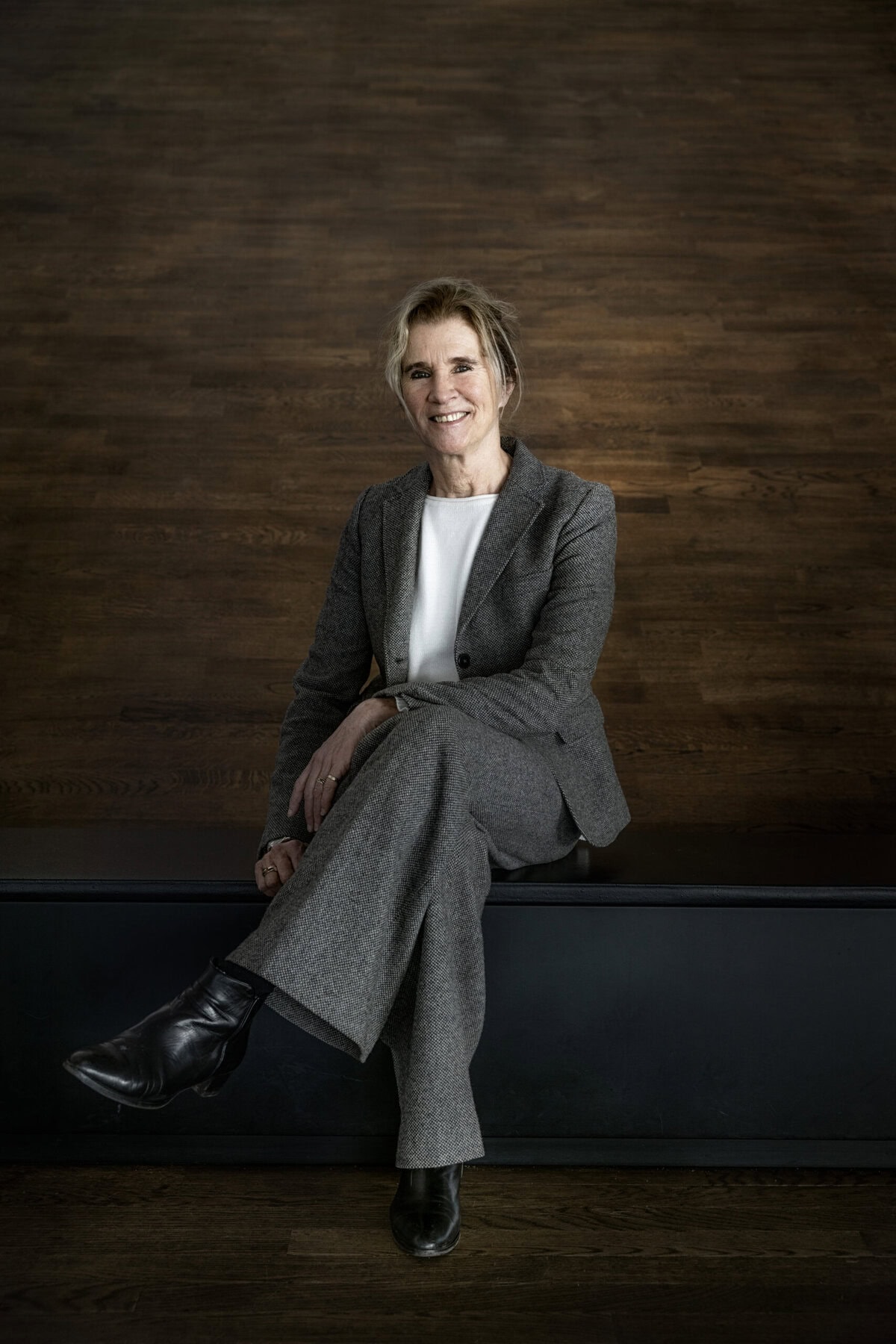 Inspirerende vrouw in zakelijke outfit zit ontspannen op bankje voor een houten achtergrond.